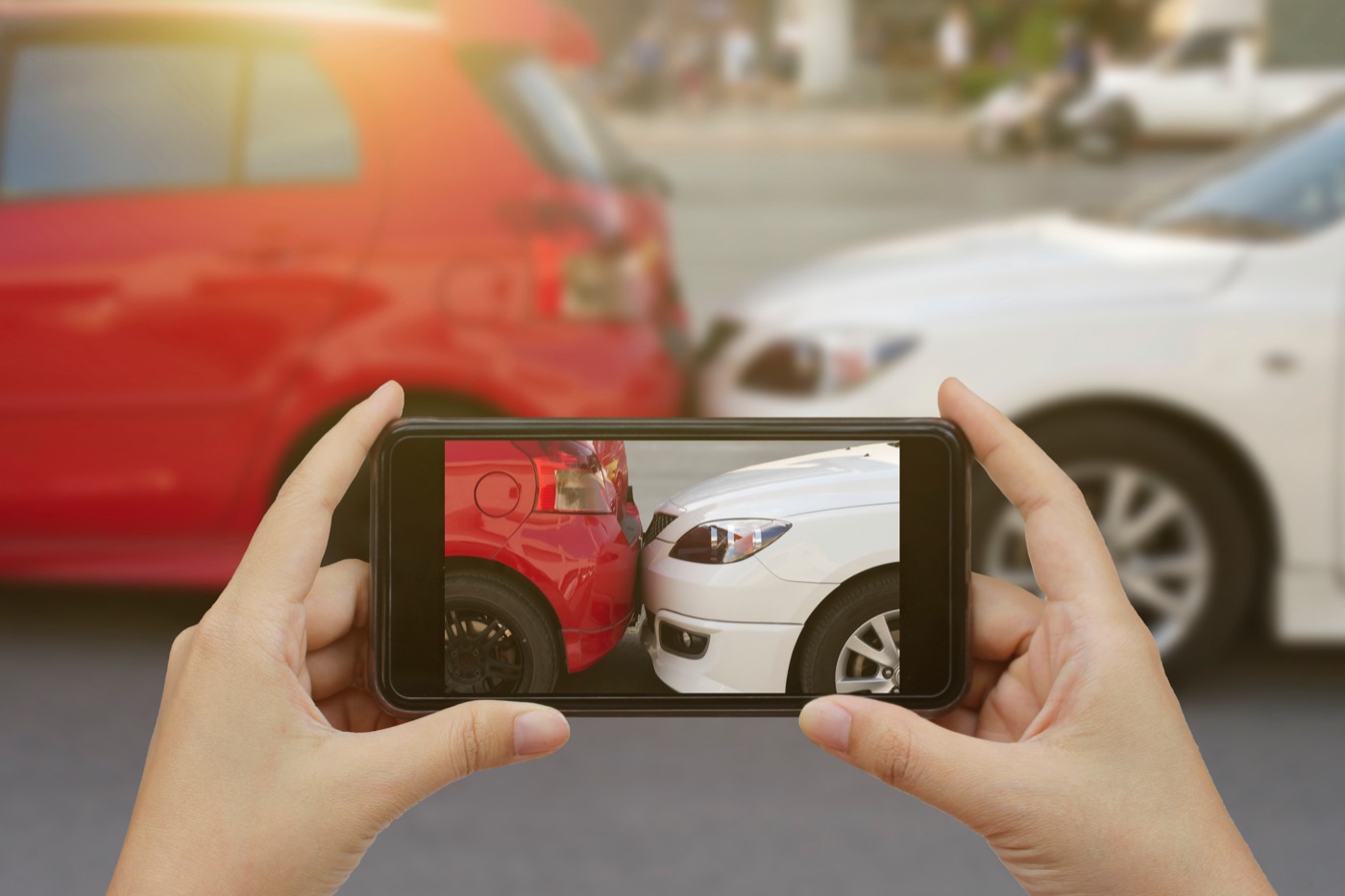 A person standing beside their car after a crash, photographing vehicle damage with their phone while emergency lights flash in the background